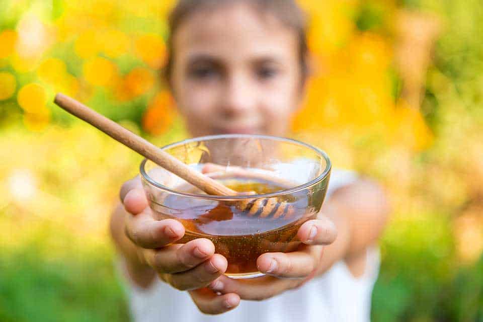 Raw honey for hay fever – child holding glass bowl of pure local raw honey with wooden dipper in sunny flower garden