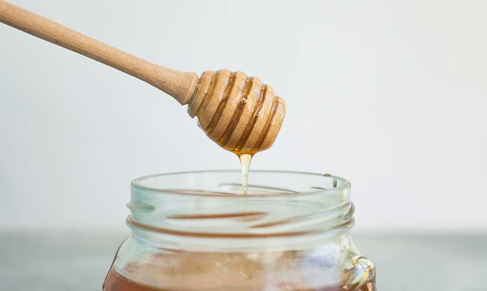 Wooden honey dipper dripping golden raw honey into a glass jar – does honey expire and shelf life of properly stored raw honey